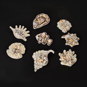 May include: A collection of eight decorative patches shaped like various seashells and marine life. Each patch is intricately embellished with white pearls, clear crystals, small tan seashells, and tiny starfish accents on a white base.