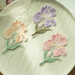 Könnte beinhalten: Drei bestickte Stoffaufnäher in Form von Irisblüten. Ein Aufnäher ist lila mit grünen Blättern, einer ist pfirsichfarben mit hellgrünen Blättern und einer ist rosa mit hellgrünen Blättern. Jede Blume weist detaillierte Stickereien auf.