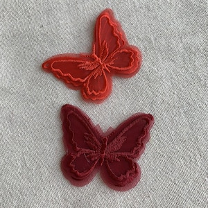 May include: Two red butterfly appliques. The butterflies are made of a sheer, layered material with embroidered details. The top butterfly is a brighter red, while the bottom one is a deeper burgundy colour. They are set against a neutral, textured background.