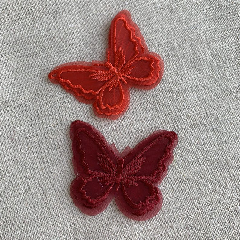 May include: Two red butterfly appliques. The butterflies are made of a sheer, layered material with embroidered details. The top butterfly is a brighter red, while the bottom one is a deeper burgundy colour. They are set against a neutral, textured background.