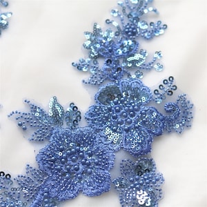 Könnte beinhalten: Eine hellblaue florale Applikation mit aufwendiger Stickerei, schimmernden Pailletten und kleinen runden Perlen. Das Design ist auf einer transparenten Netzbasis angeordnet und zeigt detaillierte Blütenformen und Blattakzente in einem einheitlichen Blauton.
