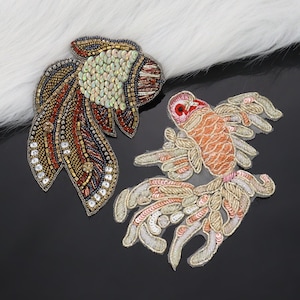 May include: Two decorative fish patches. One patch features a detailed design with sequins and beads in shades of brown, gold, and blue. The other patch is orange, pink, and gold. Both patches are on a black surface with a white fluffy background.