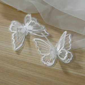 May include: Two white butterfly appliques with delicate embroidered details. The butterflies have translucent wings and are made of a sheer fabric. They are placed on a light wooden surface.