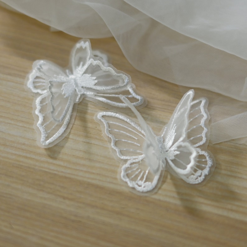 May include: Two white butterfly appliques with delicate embroidered details. The butterflies have translucent wings and are made of a sheer fabric. They are placed on a light wooden surface.