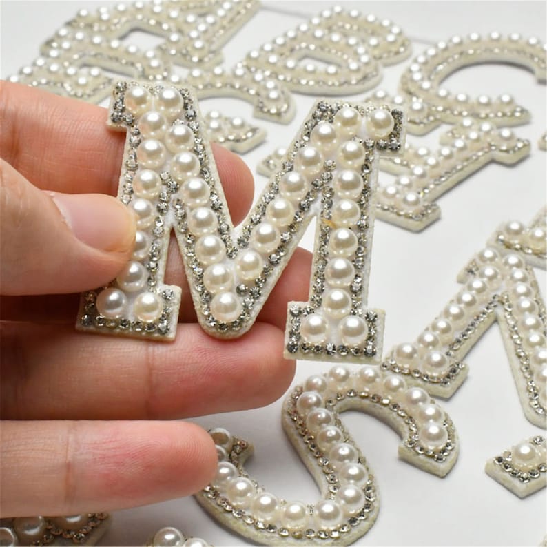 May include: A set of pearl and rhinestone alphabet letters. The letter M is being held in a hand. The letters are white with silver rhinestones and white pearls.