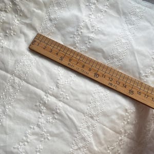 May include: White cotton fabric with a delicate embroidered pattern of small flowers and vines. The fabric is measured with a ruler marked in centimeters.