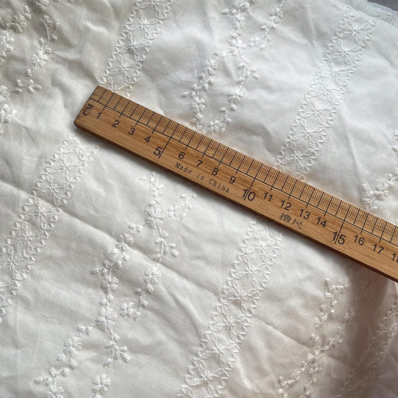 May include: White cotton fabric with a delicate embroidered pattern of small flowers and vines. The fabric is measured with a ruler marked in centimeters.