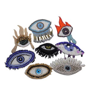 Op de afbeelding: Assortiment van kleurrijke kralen-oogpatches met verschillende ontwerpen, waaronder een vlam, een hand en een traditioneel boze oog. De patches zijn gemaakt van kralen en pailletten.