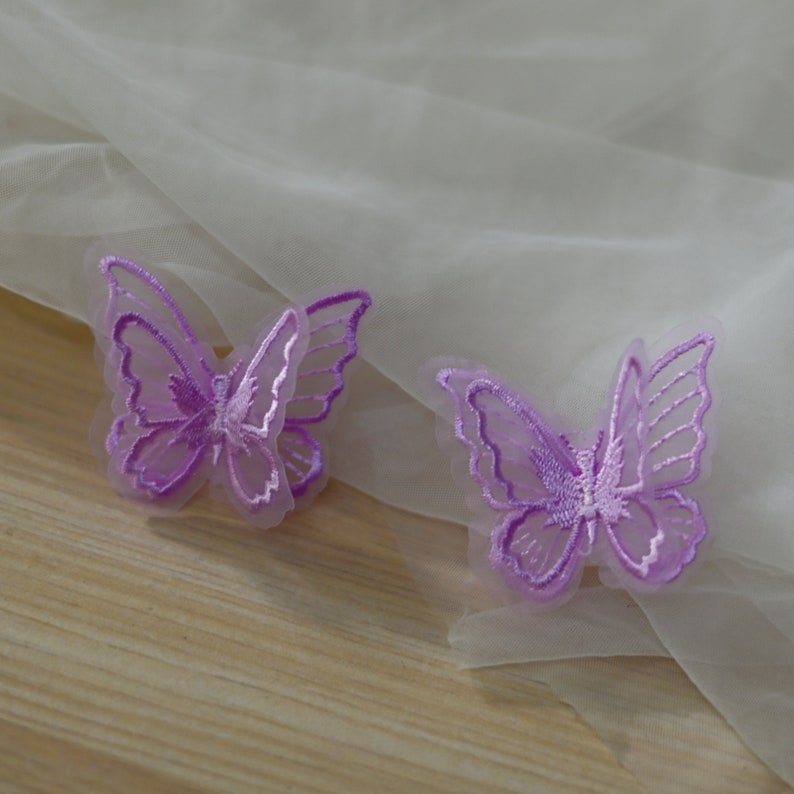 May include: Two purple butterfly appliques with embroidered details. They are set against a sheer, off-white fabric. These appliques are likely for crafting or embellishment. The butterflies have delicate wing details.