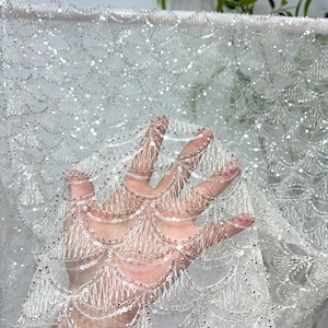 May include: Close-up of white fabric with a delicate, scalloped pattern. The fabric is embellished with shimmering sequins and beads, creating a sparkling effect. The material appears to be a sheer, lightweight tulle, ideal for bridal or formal wear.