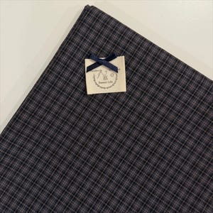 May include: A dark grey and black plaid fabric featuring a small, off-white rectangular label attached with a navy blue ribbon bow. The label displays a circular graphic with a needle, thread, and the text Sweet Life printed in black ink.