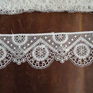 May include: A strip of white lace trim featuring a repeating geometric pattern with circular motifs and small raised dots. The lace has a scalloped bottom edge and a straight top edge, showcasing intricate thread work and openwork detailing.