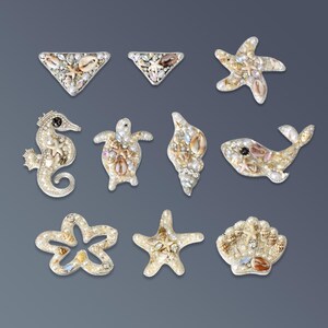 Puede incluir: Un conjunto de once parches decorativos con diversas formas y criaturas marinas, incluyendo un caballito de mar, una tortuga, una ballena, estrellas de mar y conchas. Cada pieza está adornada con pequeñas perlas blancas, cristales transparentes y conchas naturales.