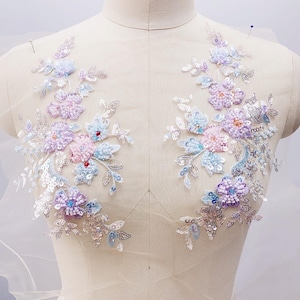 May include: Two floral appliques in pastel shades. Each features embroidered flowers in pink, purple, and blue, with silver sequins and beads. The appliques are attached to a sheer, off-white fabric, creating a delicate and ornate design, suitable for embellishment.