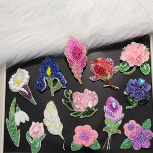 Könnte beinhalten: Eine Sammlung von zwölf verschiedenen blumen- und federförmigen Stickaufnähern mit aufwendigen Perlenarbeiten, Pailletten und Metallicfäden in leuchtenden Rosa-, Blau-, Lila-, Weiß- und Grüntönen, angeordnet auf einer dunklen Oberfläche.