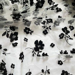 May include: White sheer fabric with black floral embroidery. The fabric has a delicate, airy feel and is perfect for creating garments or accessories.