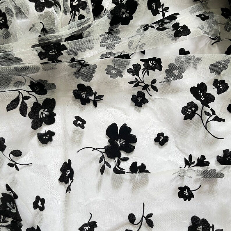 May include: White sheer fabric with black floral embroidery. The fabric has a delicate, airy feel and is perfect for creating garments or accessories.