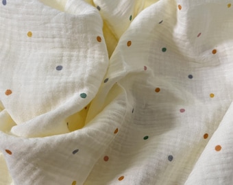 May include: Cream-coloured muslin fabric with a crinkled texture, scattered with small, colourful polka dots in shades of orange, green, blue, and pink. The fabric is draped, creating soft folds and shadows.
