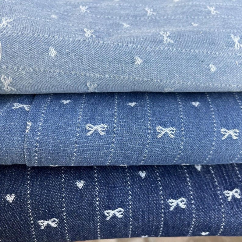 Puede incluir: Tres piezas de tela vaquera azul con patrones de lazo y coraz&oacute;n blancos. La tela est&aacute; doblada en una pila.