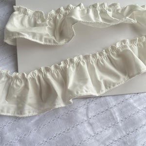 May include: Close-up of two sections of ivory-colored ruffled fabric. The fabric has a delicate, wavy edge and is likely used for trimming clothing or home decor items. The background is a plain white surface and a textured white fabric.