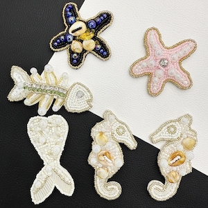 Puede incluir: Una colección de cinco parches bordados con diseños de vida marina. Se incluyen una estrella de mar azul oscuro con conchas, una estrella de mar rosa claro con cuentas, un esqueleto de pez blanco, una cola de sirena y dos caballitos de mar blancos.