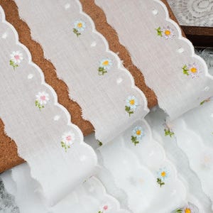 Op de afbeelding: Een witte stoffen rand met een geschulpte rand en geborduurde bloemmotieven in blauwe, roze en gele tinten. Het halfdoorzichtige materiaal is versierd met kleine witte stippen die tussen de individuele bloempatronen zijn verspreid.