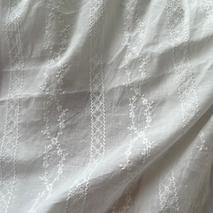 May include: White cotton fabric with a delicate embroidered pattern of small flowers and leaves. The fabric is sheer and has a soft, flowing texture.