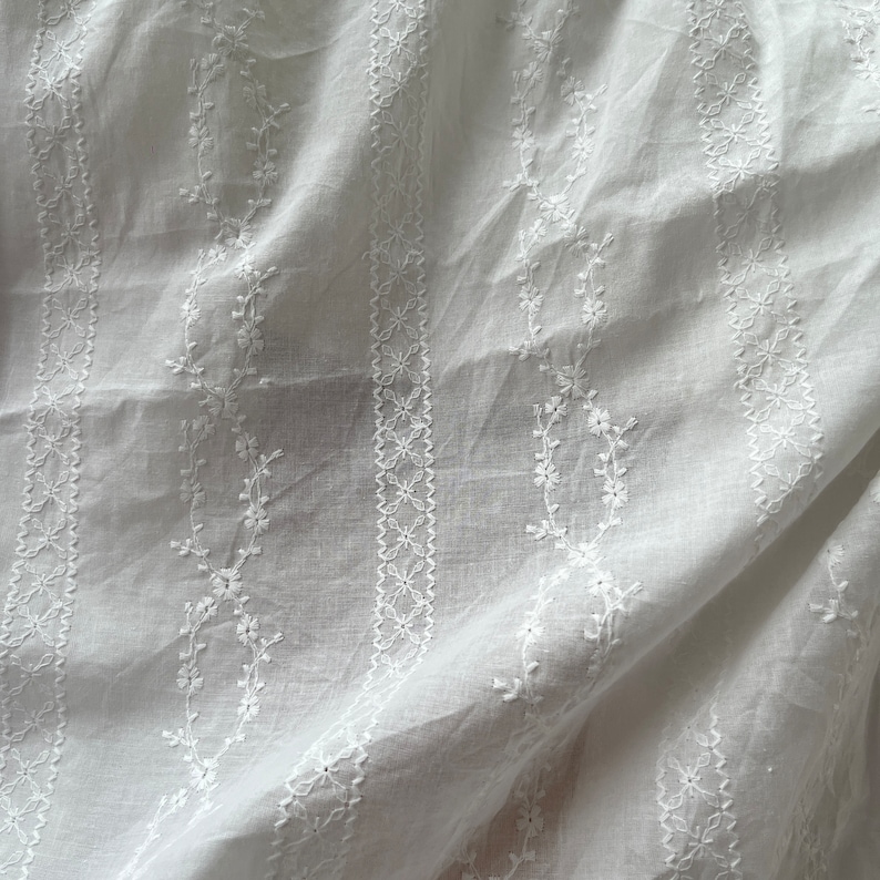 May include: White cotton fabric with a delicate embroidered pattern of small flowers and leaves. The fabric is sheer and has a soft, flowing texture.