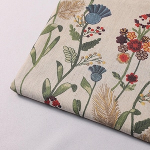 May include: A piece of off-white fabric featuring an embroidered botanical pattern. The design includes blue thistle-like flowers, red berries, yellow buds, and various green leaves and stems, along with textured golden-brown wheat-like stalks.