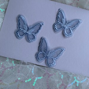 May include: Three pale blue butterfly appliques with intricate embroidery. They are displayed on a pale pink surface, with a sheer, sequined fabric in the foreground. These appliques are suitable for various craft projects.