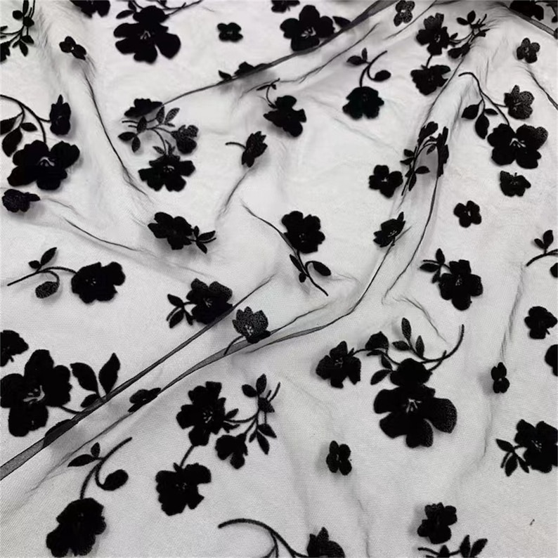 May include: Black sheer fabric with black velvet floral embroidery. The fabric has a delicate, airy feel and is perfect for creating elegant garments or accessories.