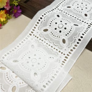 May include: A white cotton fabric trim featuring an intricate eyelet embroidery pattern. The design consists of repeating square motifs with circular centers and oval cutouts, bordered by delicate scalloped edges and small circular perforations.