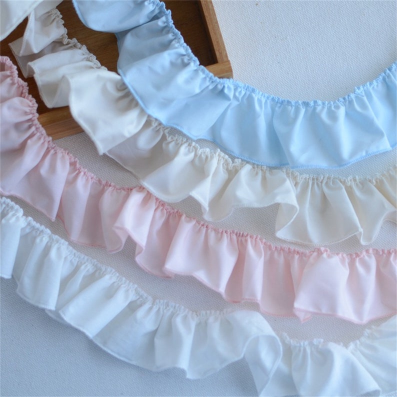 May include: Close-up of several rows of ruffled fabric trim in pastel colors. The colors include light blue, cream, pink, and white. The fabric has a delicate, airy appearance, with soft folds and a subtle sheen. Ideal for sewing and craft projects.