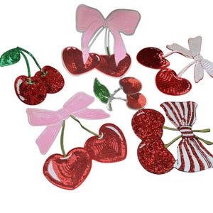 Puede incluir: Una colección de parches de cerezas y lazos con lentejuelas. Las cerezas son principalmente rojas con tallos verdes, mientras que los lazos son rosas y blancos. Estos parches probablemente se utilizan para adornar ropa o accesorios.