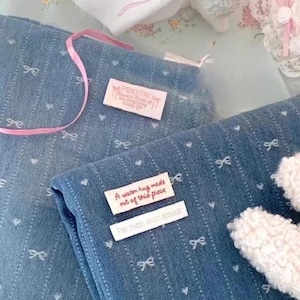 Puede incluir: Dos etiquetas de tela sobre una tela vaquera azul con un patr&oacute;n de lazo blanco. La etiqueta superior es rosa y dice "FORYOU". La etiqueta inferior es blanca y dice "A warm hug made out of this piece" y "Felt with love and amour".