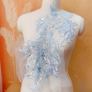 May include: A light blue iridescent sequin applique in the shape of a fish tail. The applique is on a white mesh background.