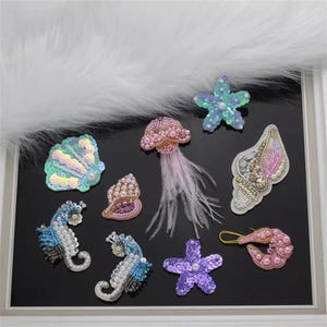May include: Assortment of colourful, beaded, and sequined iron-on patches featuring sea creatures, including a starfish, seashell, jellyfish, and seahorse.