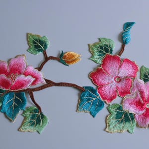 May include: Embroidered floral applique featuring pink flowers with red centers, green and teal leaves, and a brown stem. The flowers have detailed stitching and a gradient effect, creating a realistic look. The applique is likely for clothing or craft projects.