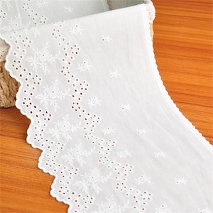 Op de afbeelding: Een witte katoenen stoffen rand met een ingewikkeld broderie anglaise patroon met bloemmotieven en geschulpte randen. Het textiel toont een herhalend ontwerp van kleine bloemen en cirkelvormige uitsparingen, afgewerkt met een fijne schulprand.