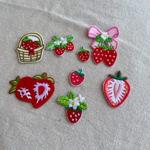 Op de afbeelding: Een verzameling geborduurde aardbeienpatches in verschillende ontwerpen. Inclusief een mand met aardbeien, aardbeien met bladeren en bloemen, en individuele aardbeien. De patches zijn rood, groen, wit en roze.