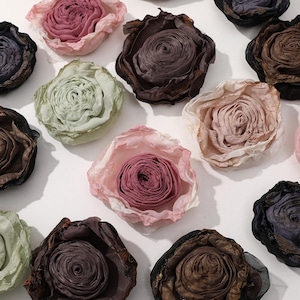 Può includere: Una vista dall'alto mostra diversi abbellimenti floreali in tessuto di vari colori. I fiori variano dal rosa chiaro al marrone scuro, alcuni con un design a petali stratificati. La composizione è su sfondo bianco.