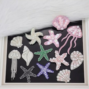 May include: A collection of 12 pearl and sequin embellished iron-on patches in the shape of seashells and starfish. The patches are pink, white, green, and blue.