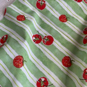May include: A fabric featuring a repeating pattern of bright red tomatoes with green stems scattered across a background of alternating light green and white vertical stripes. The tomatoes appear in various orientations, some whole and some halved.