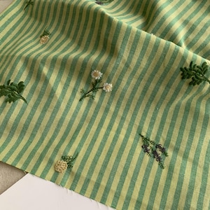 Pode incluir: Um tecido dobrado com um padrão listrado em verde claro e amarelo. O tecido é adornado com desenhos florais bordados em branco, verde e roxo. O bordado adiciona um toque delicado ao design geral do tecido.