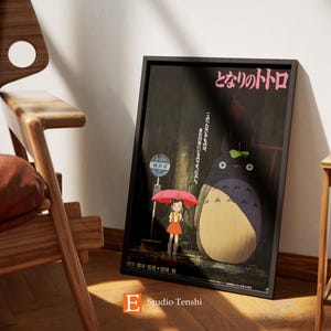 Puede incluir: Un p&oacute;ster enmarcado en negro con el texto japon&eacute;s "となりのトトロ" (Totoro) y una ilustraci&oacute;n de una gran criatura peluda gris con una ni&ntilde;a peque&ntilde;a sosteniendo una sombrilla rosa bajo la lluvia.