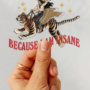 Puede incluir: Una pegatina troquelada que muestra a una persona con sombrero de vaquero montando un tigre alado. El texto I WILL SUCCEED BECAUSE I AM INSANE está impreso en letras rojas arqueadas sobre y bajo la ilustración, decorada con pequeñas estrellas amarillas.