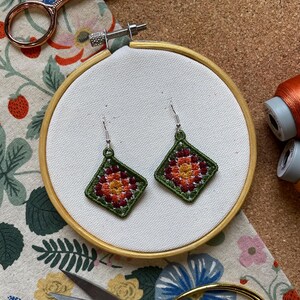 以下が含まれることがあります： 手作りの刺繍イヤリングのペア。各イヤリングは、オレンジ、赤、黄色の色合いの、中央に花の模様がある緑色のひし形のフレームが特徴です。イヤリングは、木製の刺繍枠の中の白い布に展示されています。