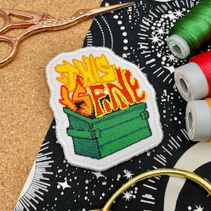 Peut inclure: Écusson brodé représentant une poubelle verte en feu avec les mots "THIS IS FINE" en jaune et rouge. L'écusson est sur fond blanc. Des fournitures de couture, dont du fil et des ciseaux, sont en arrière-plan.