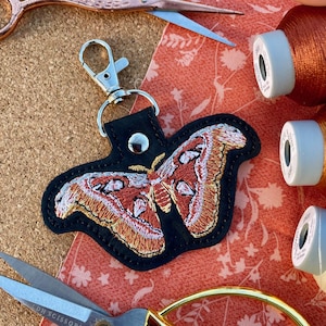 May include: A black leather keychain with a detailed embroidered moth in shades of orange, red, and white. The keychain has a silver metal clasp.