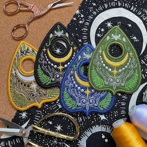 Könnte beinhalten: Vier gestickte Patches in Form einer klassischen Ouija-Planchette. Die Patches sind in Gold, Grün, Blau und Schwarz mit weißen Akzenten. Die Patches befinden sich auf einem schwarzen Stoff mit weißen Sternen.